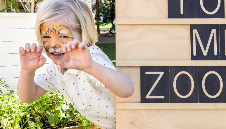 Happy child peaking around a sign with tiger face paint at a Kids Zoo Party.