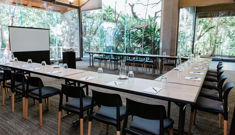 Corporate function area with U-shaped tables. Large floor to ceiling windows allowing natural light to stream in through green leafy trees.