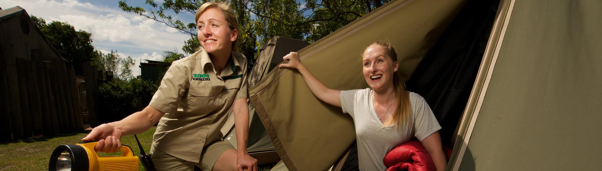 A female zoo worker crouching and holding a torch while another woman looks out of a tent door flap.