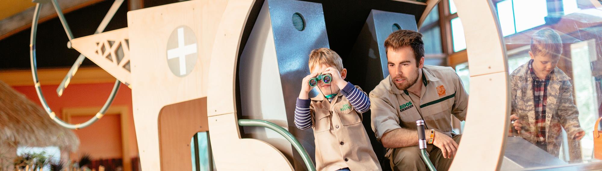 Child looking through binoculars with safari guide playing in a wooden helicopter in Ranger Kids indoor play centre.  