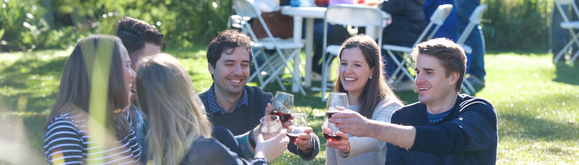Group of six young adults sitting on a picnic rug, in the gardens of Healesville Sanctuary, making a toast with red wine at Wine and Wildlife.