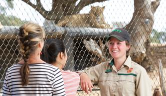 Smiling Lion Keeper chatting to two young women at the Lion Presentation. Lion enclosure in the background.