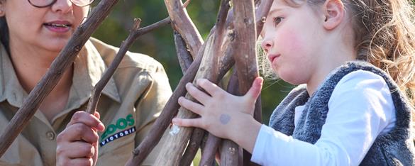 Primary aged student holding the tops of a handful of medium sized sticks. A Zoos Victoria staff member is beside the student, carefully adding a stick to her collection.  