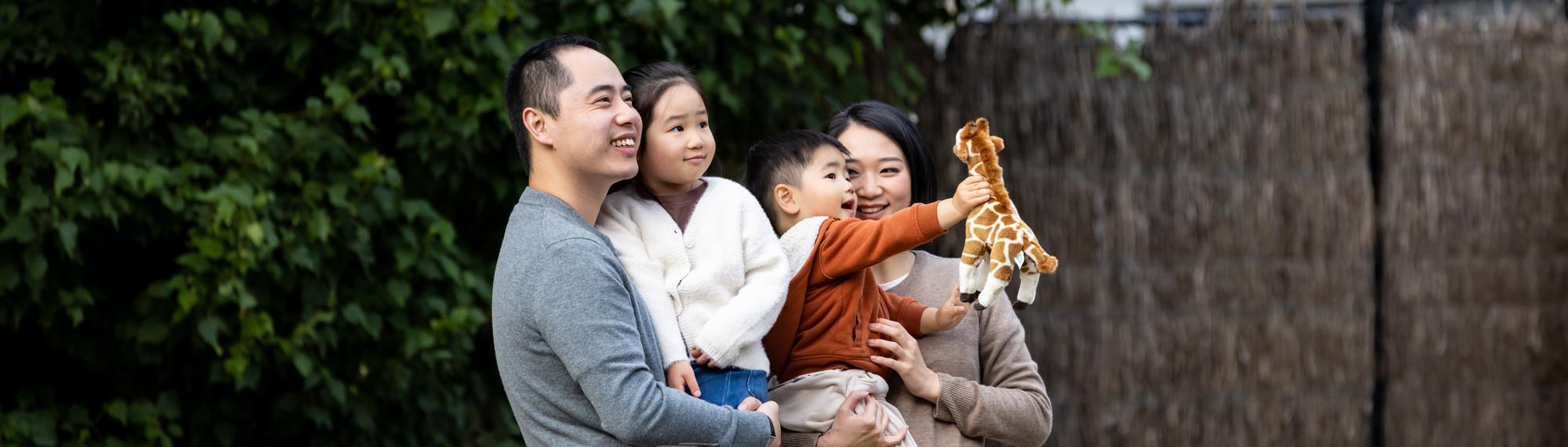 Family of two kids looking at giraffe and smiling. Young boy is holding out the giraffe plush toys