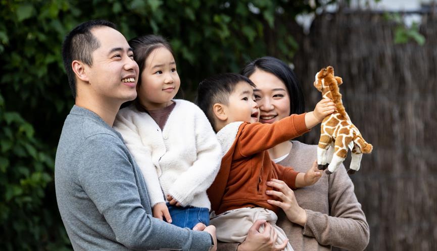 Family of two kids looking at giraffe and smiling. Young boy is holding out the giraffe plush toys