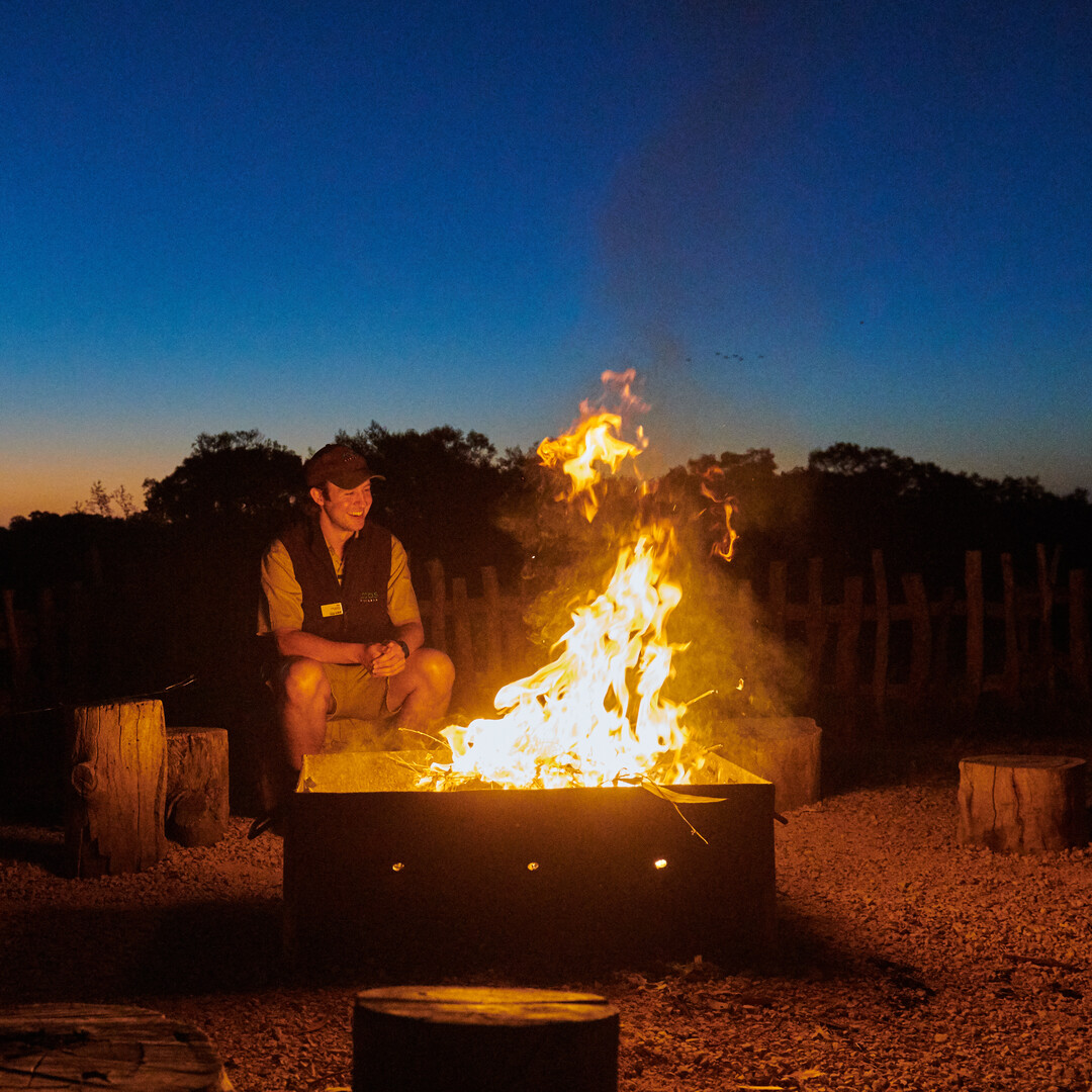 A man sits behind a camp fire at dusk. The sky is dark blue.