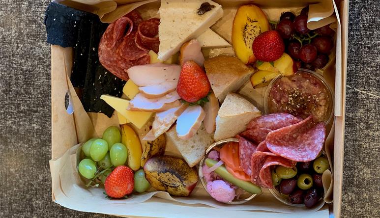 Contained in a cardboard box, seen from above: Grapes, Strawberries, Cherries, Olives, Dip, Sliced Salami, Chicken, Bread, Cheese and Mango.