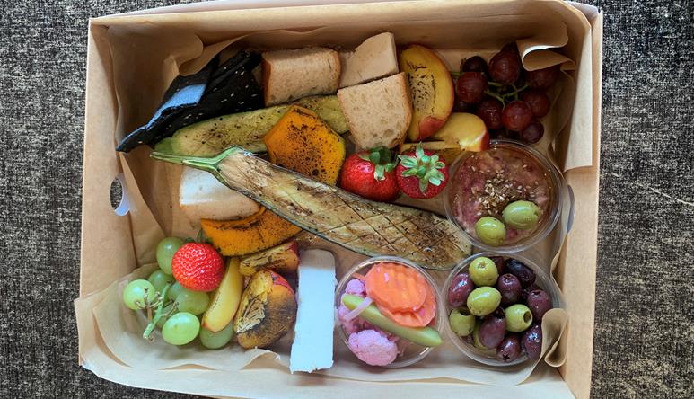 Contained in a cardboard hamper, seen from above: Cherries, Grapes, Strawberries, Olives, Dip, Sliced Bread, Mango, Pumpkin, Eggplant, and Brie Cheese.