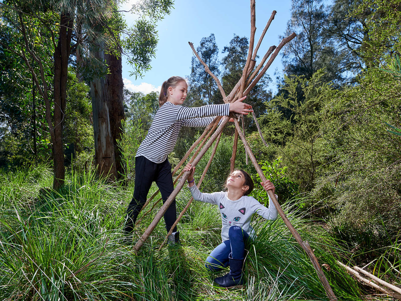 Two young girls assemble large branches to build a gunyah