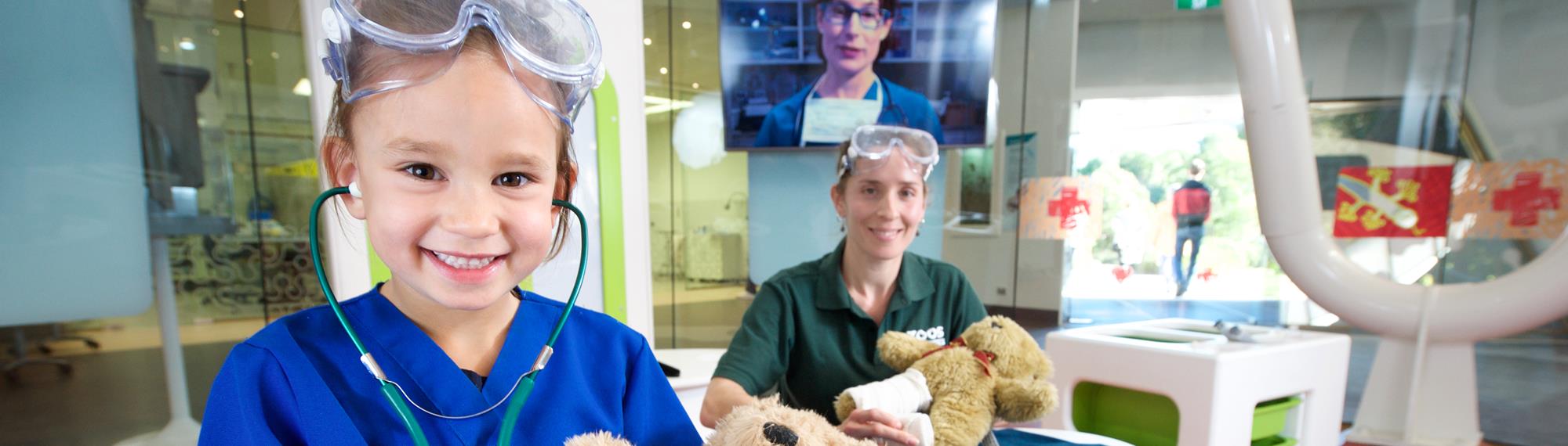 A young guest, happily playing veteranarian with her plush bear, accompanied by a volunteer behind her and both smiling to the camera, in the Australian Wildlife Health Centre.