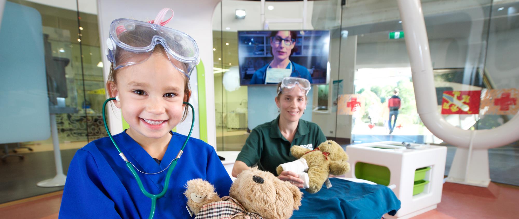 A young guest, happily playing veteranarian with her plush bear, accompanied by a volunteer behind her and both smiling to the camera, in the Australian Wildlife Health Centre.