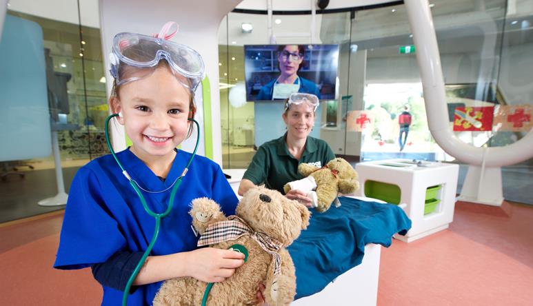 A young guest, happily playing veteranarian with her plush bear, accompanied by a volunteer behind her and both smiling to the camera.