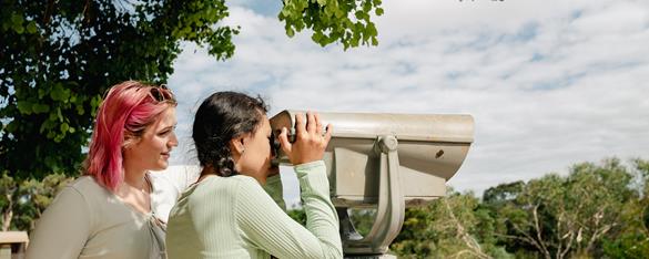 Two guests at a binocular view-post, with one looking through it toward shrubbery to the right.