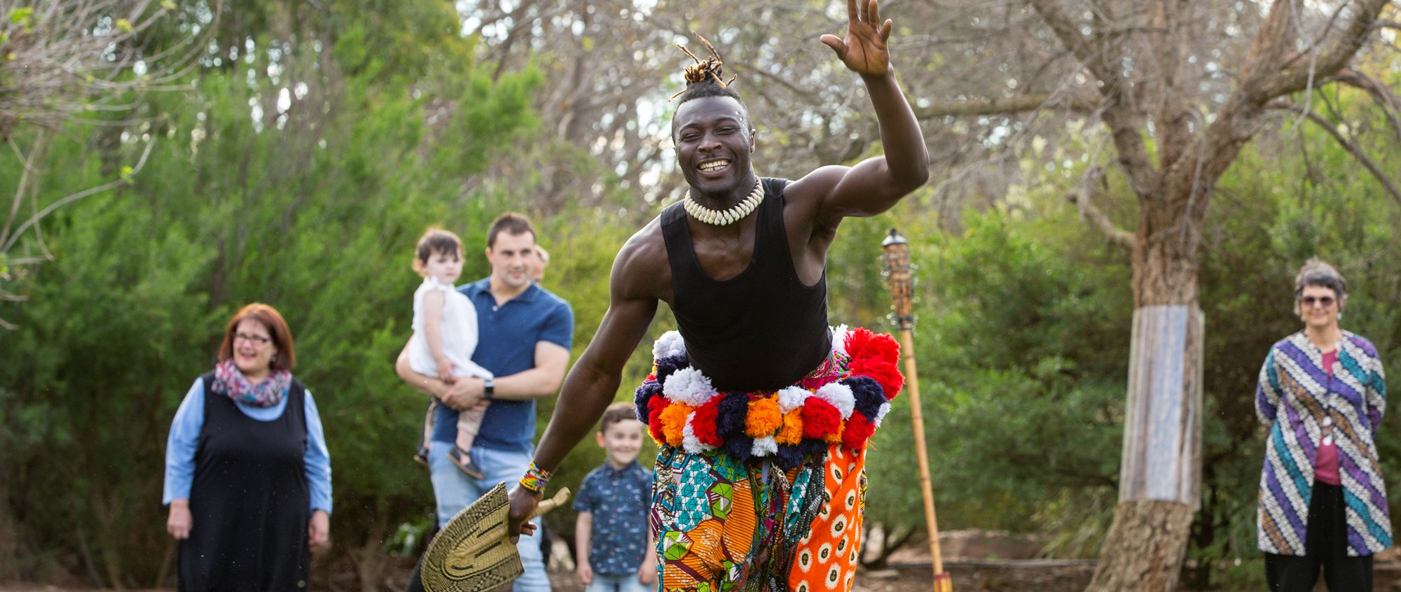 An African dancer, looking to the camera with a spade in his right hand, while five guests watch on from behind.