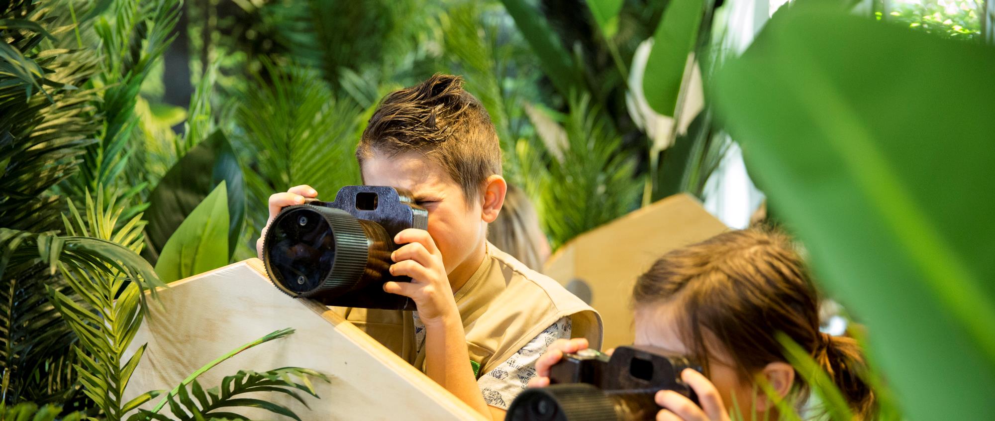 Two young guests, dressed as keepers and holding cameras, ready to take photos within a jungle setting.