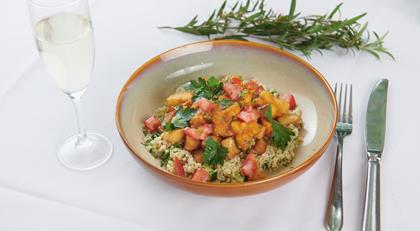 A dish of chicken, potato, rice and tomato, in a bowl on a white-clothed table, surrounded by a glass of champagne, a leafy branch, a knife and fork.